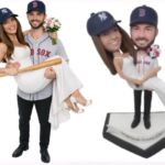 Playful wedding cake toppers featuring a couple in baseball theme: groom in a Red Sox jersey carries a bride in a white dress with a bouquet; both wear baseball caps. A second bobblehead-style figure stands on a diamond base.