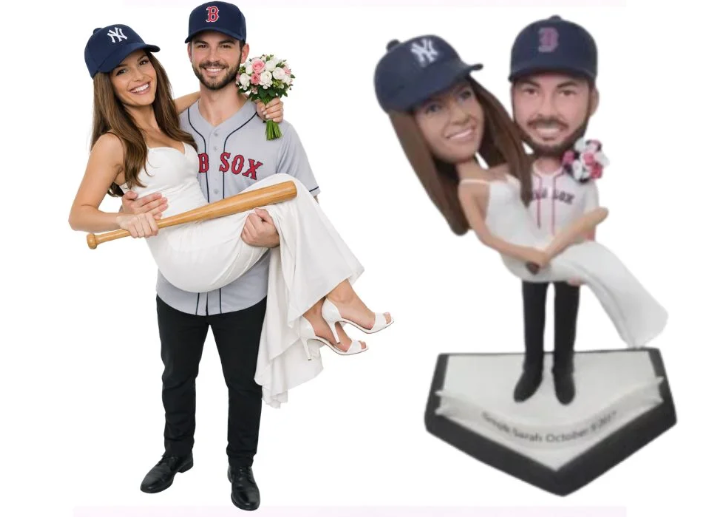 Playful wedding cake toppers featuring a couple in baseball theme: groom in a Red Sox jersey carries a bride in a white dress with a bouquet; both wear baseball caps. A second bobblehead-style figure stands on a diamond base.