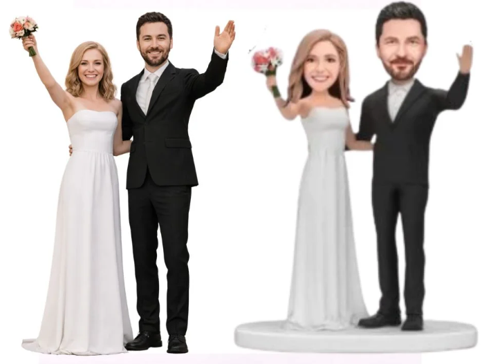 Two wedding cake toppers: a couple on the left with a bride in a white gown and groom in a black suit raising their arms in celebration; another couple on a pedestal—the bride in white and the groom in black suit—also waving.