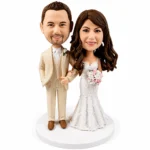 Wedding cake topper bobbleheads: groom in a beige suit and bride in a lace white gown with a bouquet on a white base.