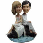 Wedding cake topper figurines: a bride and groom sitting on a rock above water, smiling as a couple.