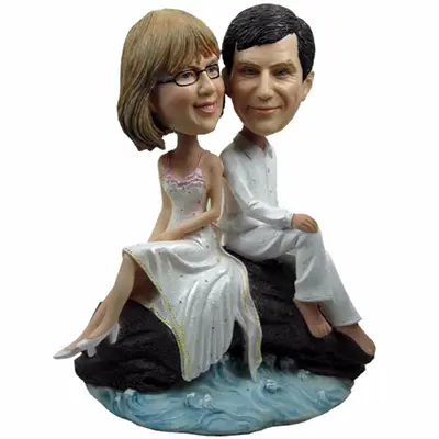 Wedding cake topper figurines: a bride and groom sitting on a rock above water, smiling as a couple.