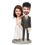 Bobblehead-style wedding couple figurine: bride in a white dress and the groom in a gray suit, standing on a circular base with bouquet in hand