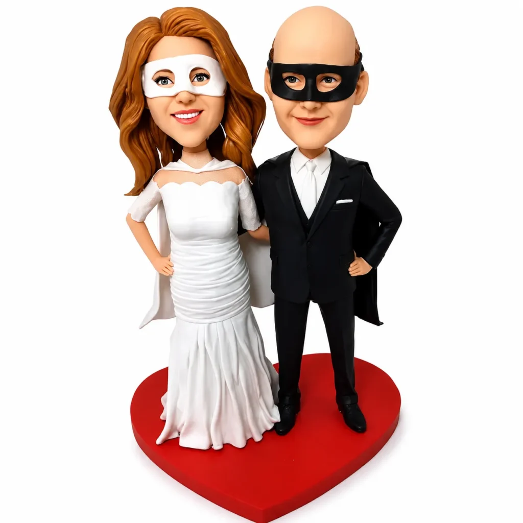 Two wedding cake toppers—a bride in a white gown and a groom in a black suit—stand side by side on a red heart-shaped base, smiling at the camera.