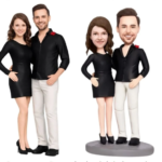 Two couples in black outfits posing together; real couple on the left and their caricature figurines on a pedestal on the right.