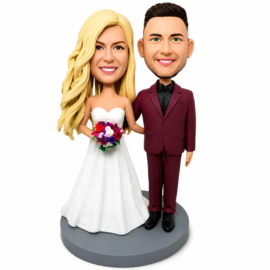 Bobblehead wedding couple: bride in a white strapless gown with a colorful bouquet and the groom in a burgundy suit, both smiling on a gray base.