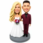 Bobblehead wedding couple: bride in a white strapless gown with a colorful bouquet and the groom in a burgundy suit, both smiling on a gray base.