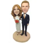 Bobblehead wedding cake topper featuring a bride in a white dress with bouquet and a groom in a dark suit standing on a beige base