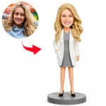 Bobblehead figure of a smiling blonde woman in a gray dress and white blazer on a round gray base; inset photo shows the same woman in a hoodie, with a red arrow linking the photo to the figure.