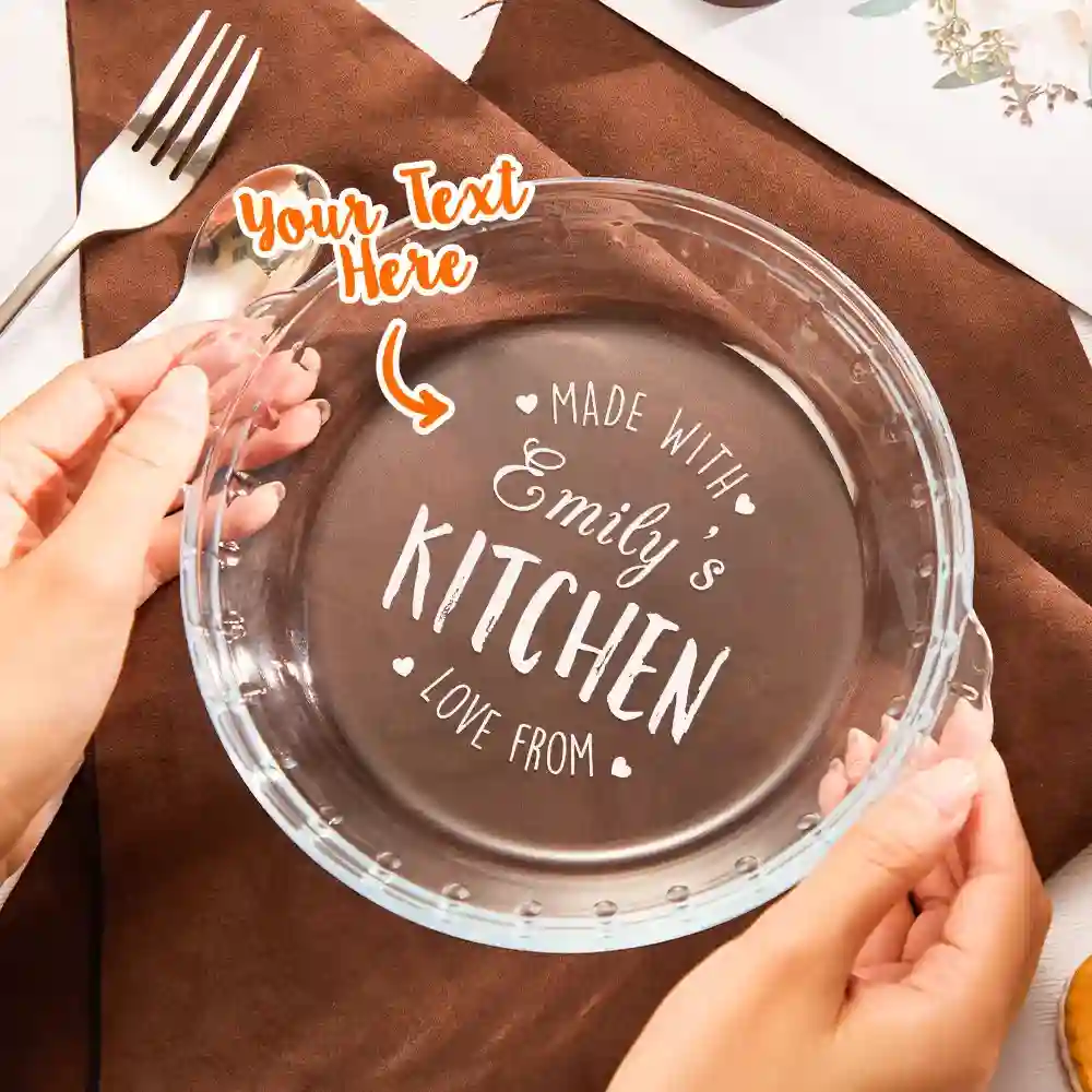 Round glass plate with customizable engraving: 'Made with Emily's Kitchen, Love From' in white script; a hand holds the edge, with a brown napkin beneath.