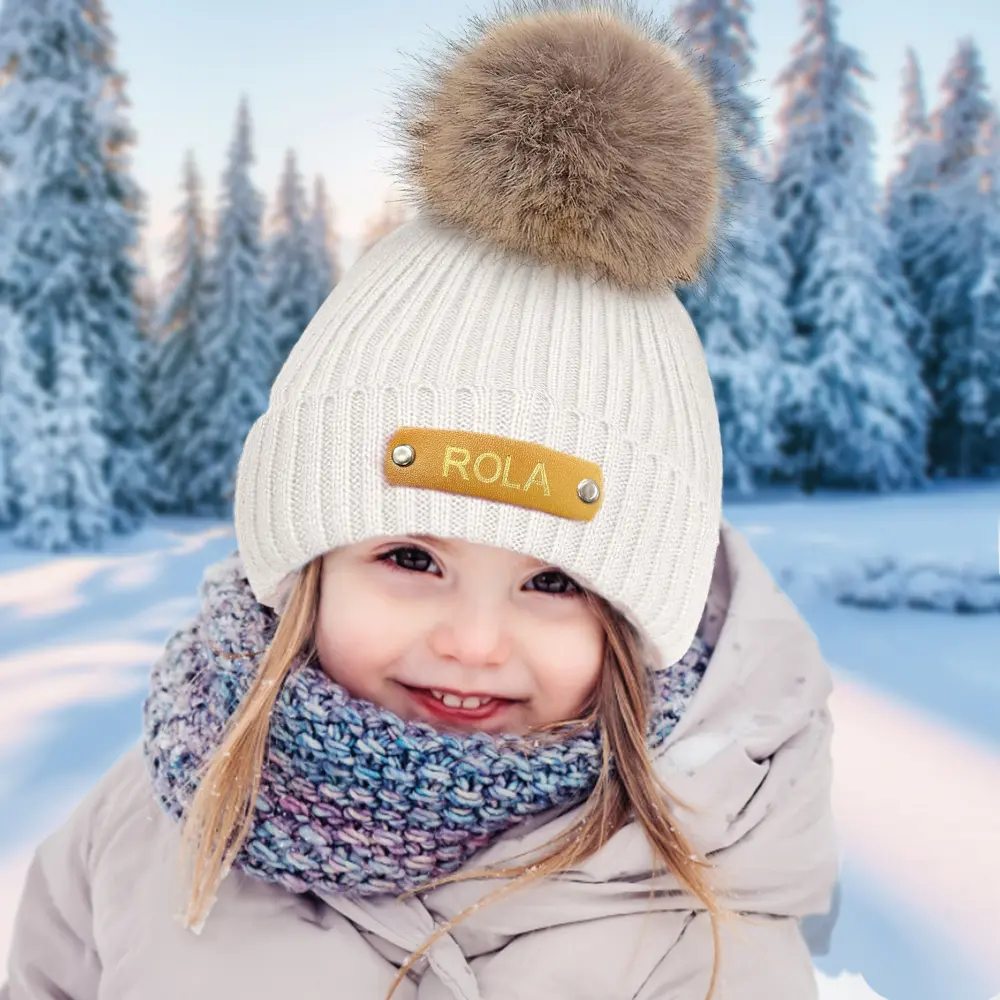 Smiling child in a white knit hat with a fluffy pom-pom, multicolored scarf, and light coat in a snowy forest backdrop.