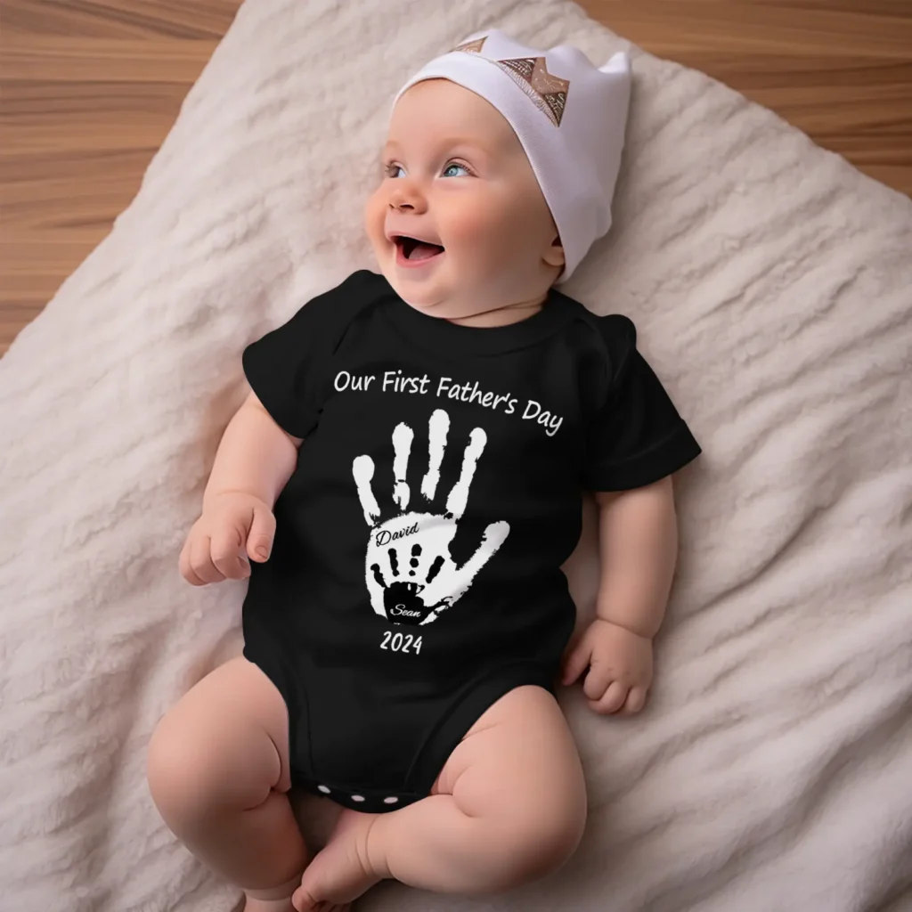 Smiling baby wearing a black onesie with a white handprint and 'Our First Father's Day' text, lying on a fluffy cream blanket.
