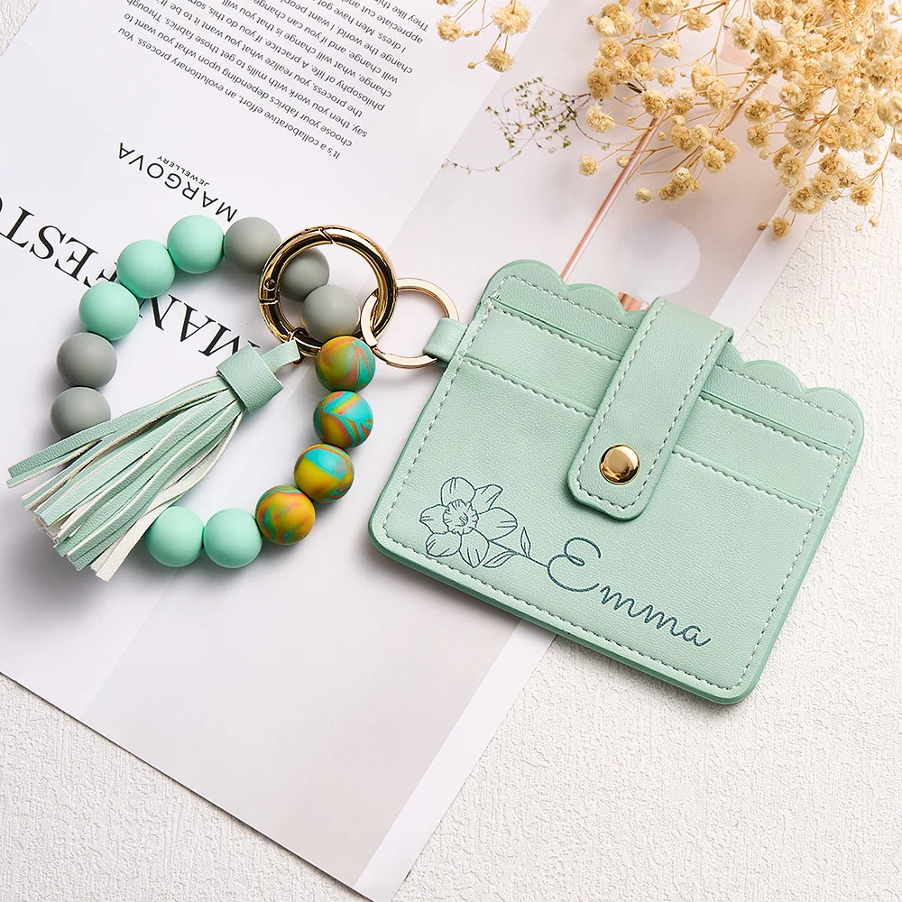 Mint-green cardholder with floral engraving and 'Emma' monogram, attached to a beaded tassel keychain with pastel teal and multicolor beads.