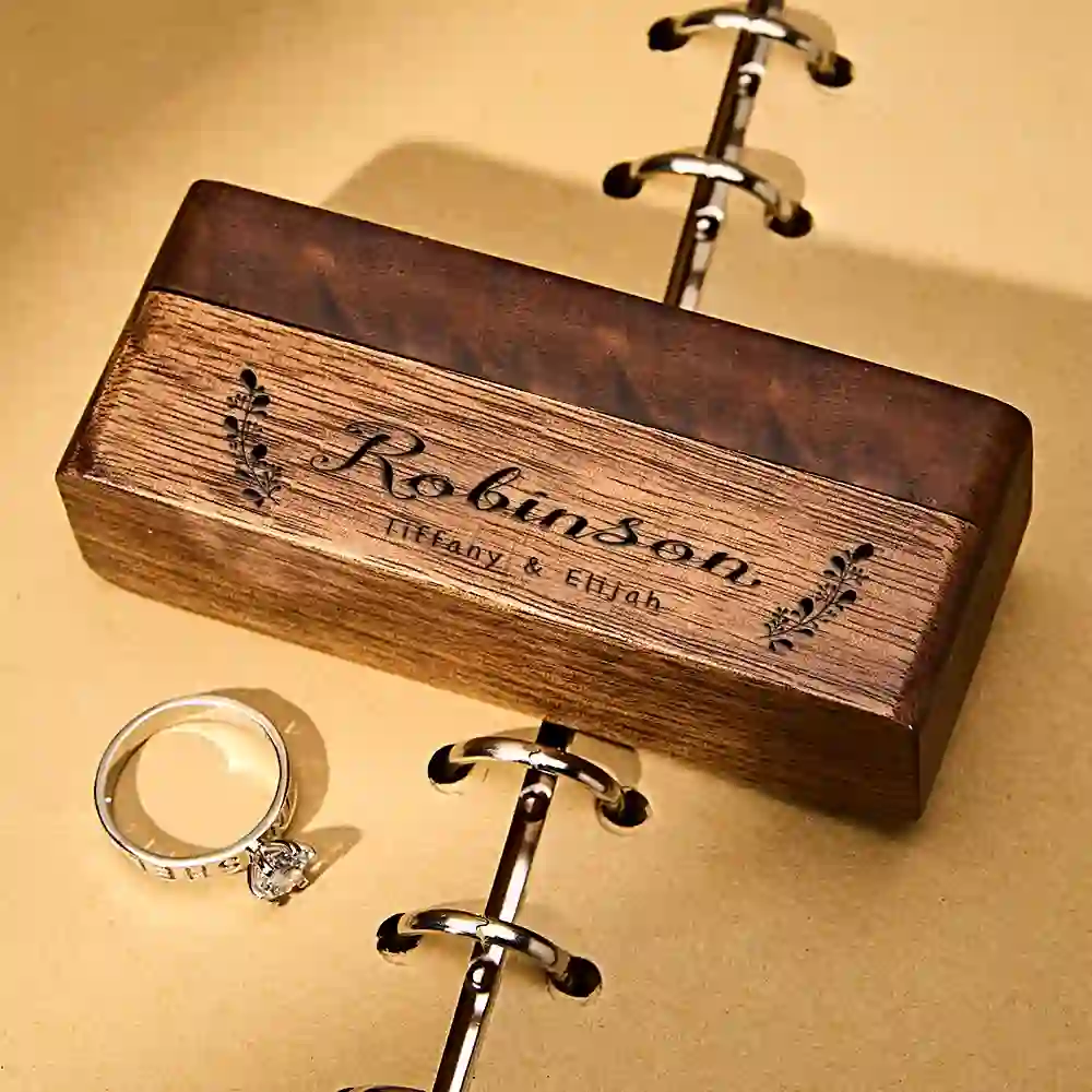 Engagement ring with a diamond next to a wooden engraved ring box resting on a gold binder chain.