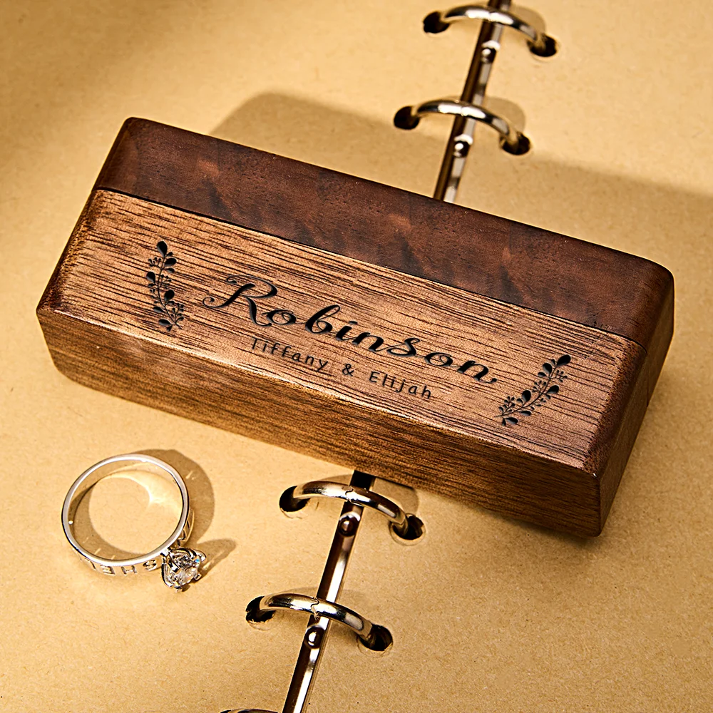 Engraved wooden ring box with metal binder rings and a diamond engagement ring beside it on a tan background.
