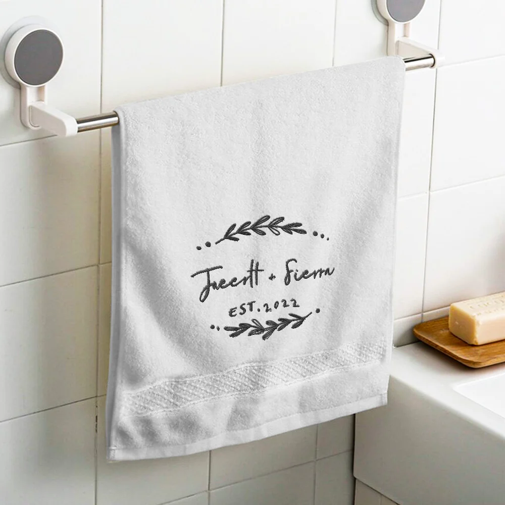 White towel hanging on a chrome bar with black botanical embroidery and 'EST. 2022' text in a white-tiled bathroom setting.