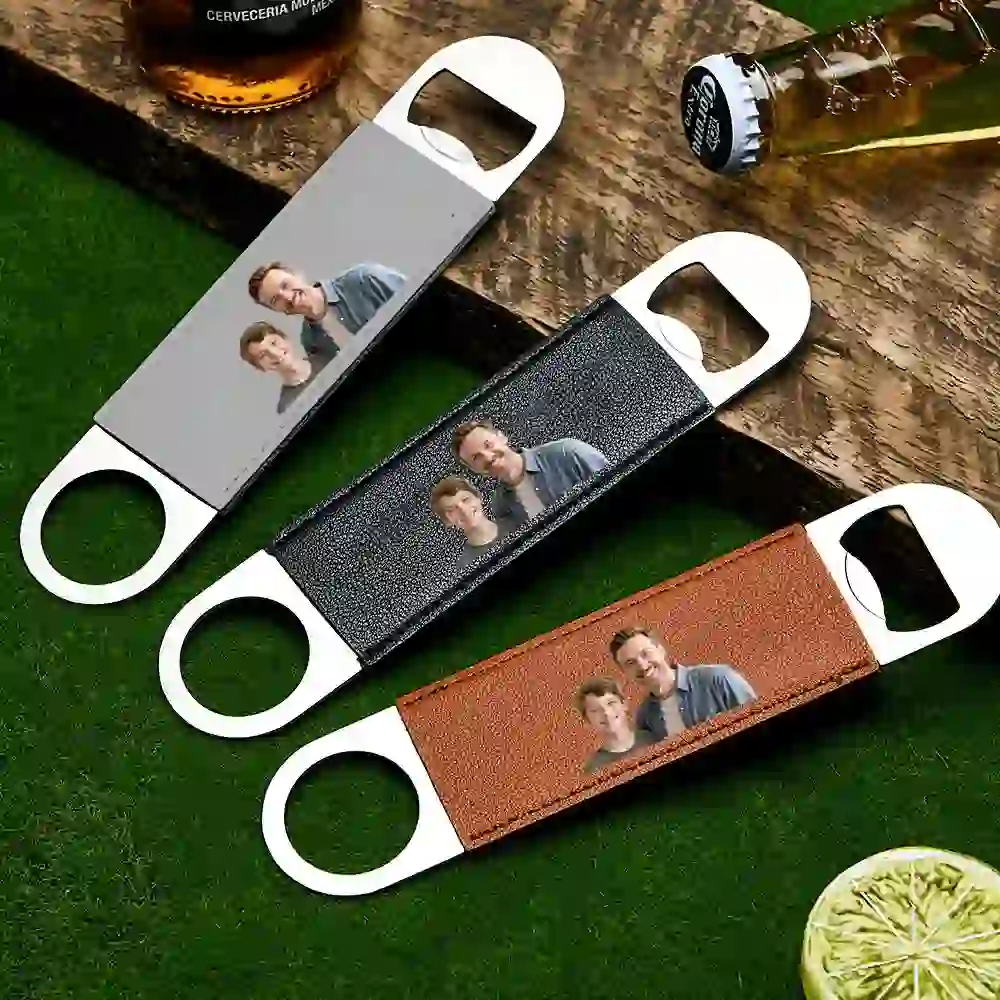 Leather strap bottle openers with a printed photo of a couple, laid on wood and grass surfaces.
