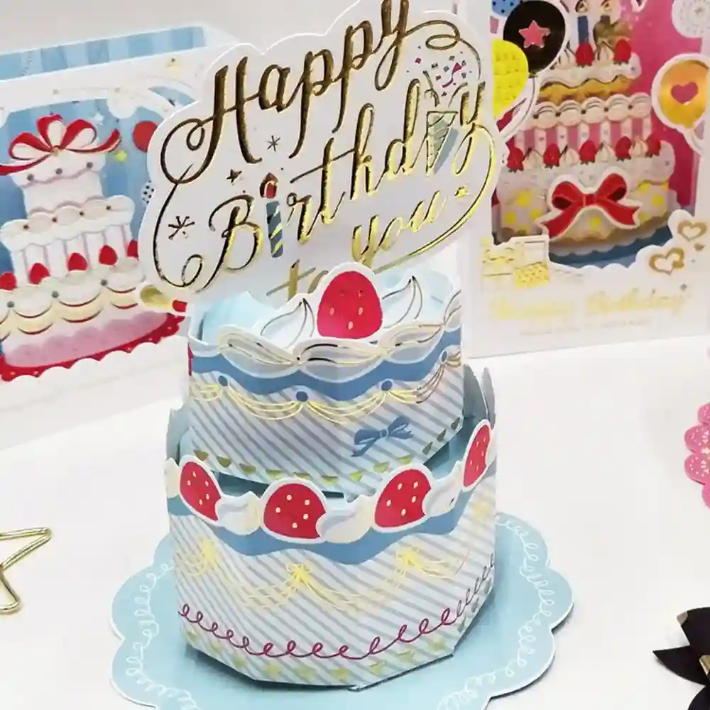 Three-tier birthday cake pop-up card with a gold 'Happy Birthday to You' topper on a blue and white cake, set on a decorative base.