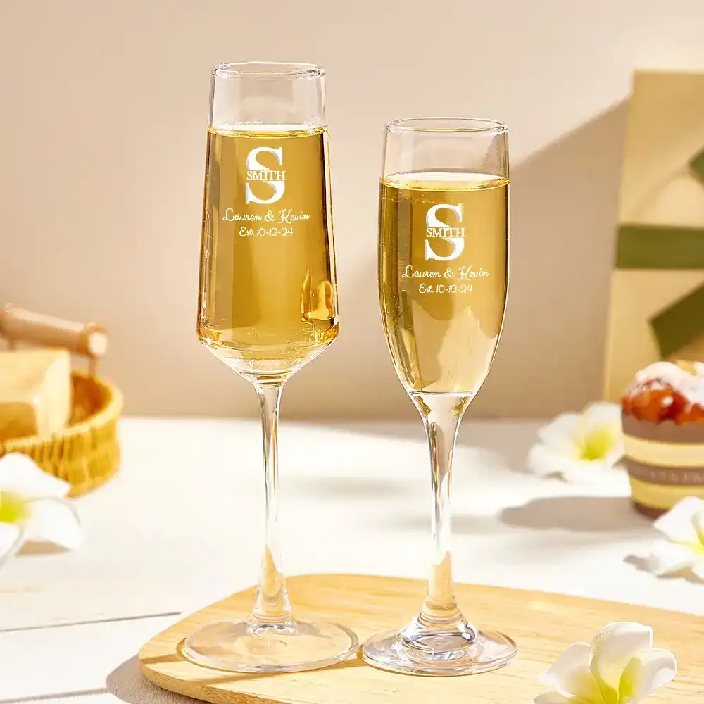 Two personalized champagne flutes with gold liquid on a round wooden board, engraved with a monogram and names for a wedding toast.