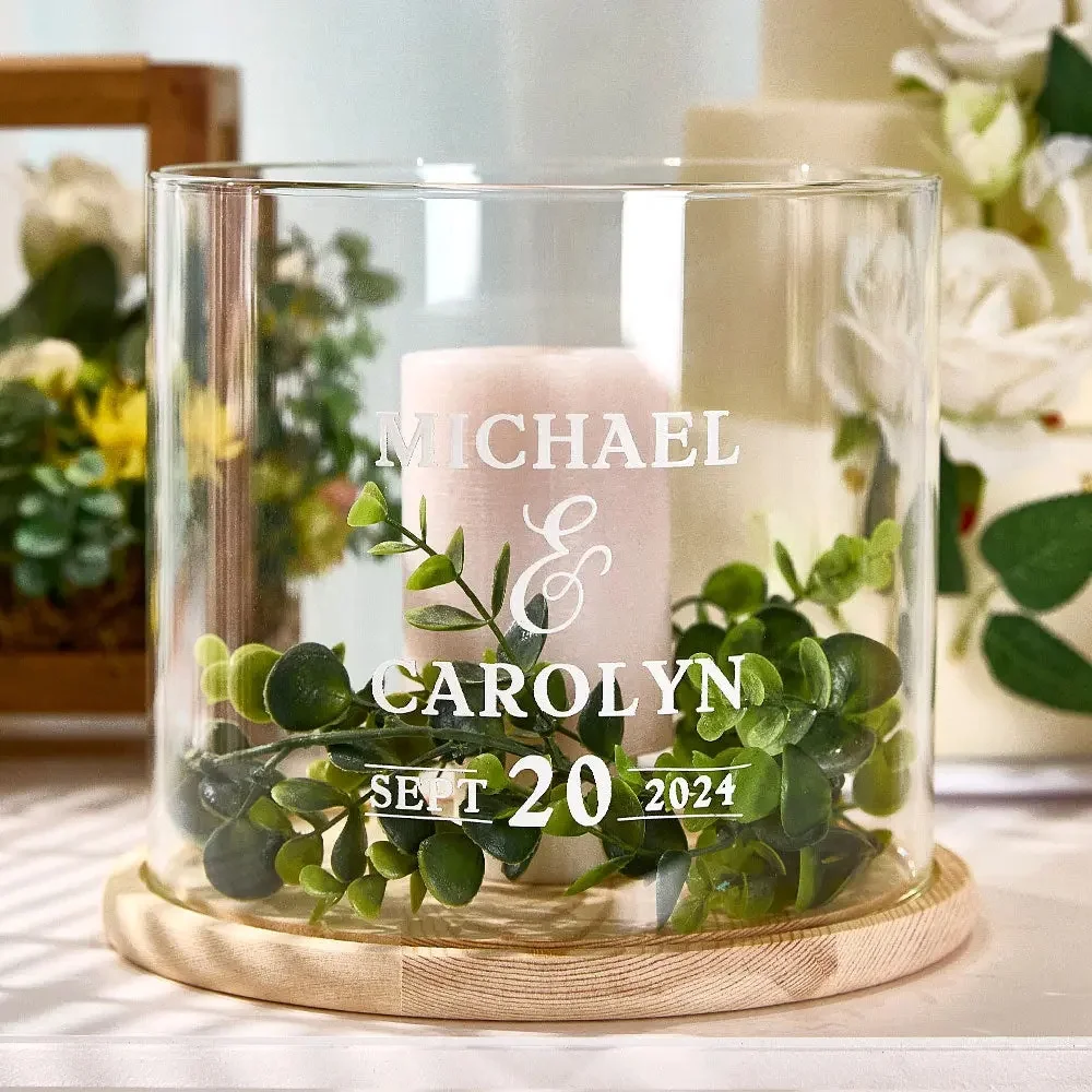 Glass hurricane vase with a pink candle and greenery, engraved 'MICHAEL & CAROLYN' and 'SEPT 20 2024'