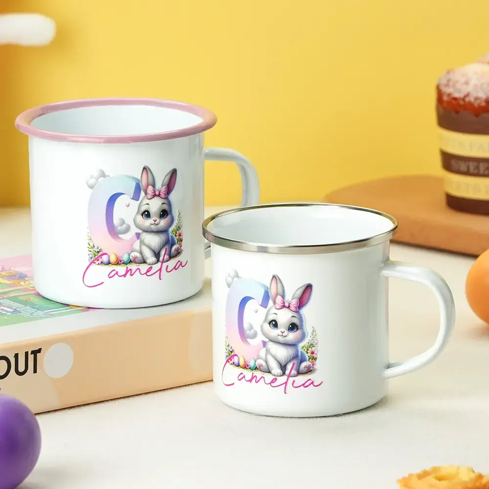 Pair of white enamel mugs with a cute bunny illustration and pink script text reading 'Camelia' on a bright yellow backdrop.