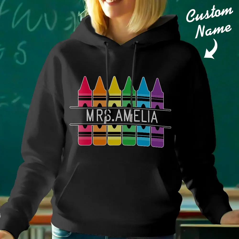 Black hoodie showing rainbow crayons arranged in two rows around the centered text 'MRS. AMELIA'; the design is customizable with a different name.