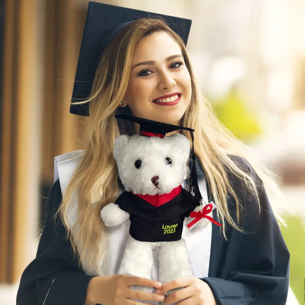 Smiling graduate in a cap and gown holds a white teddy bear wearing a mini graduation cap embroidered 'Lauren 2023'.