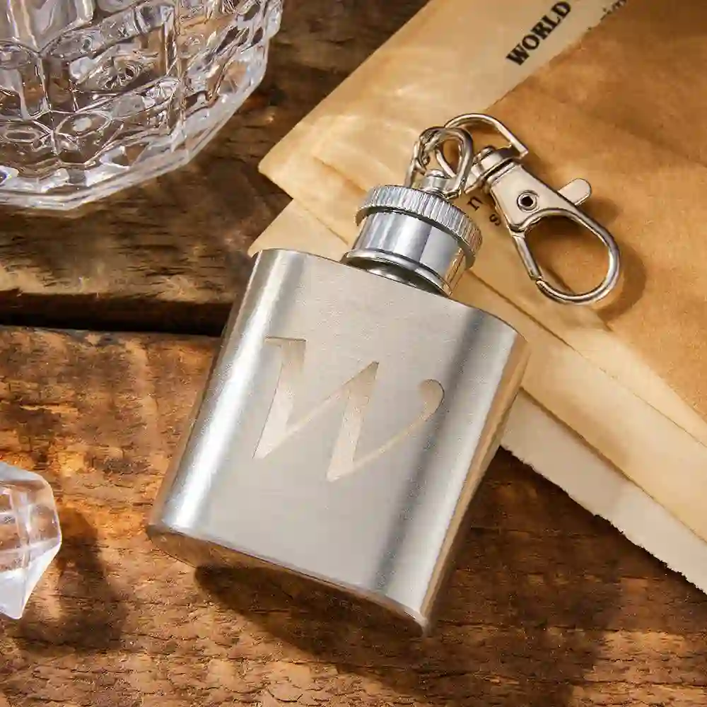 Stainless steel hip flask with a screw cap and attached keyring clip on a wooden surface.