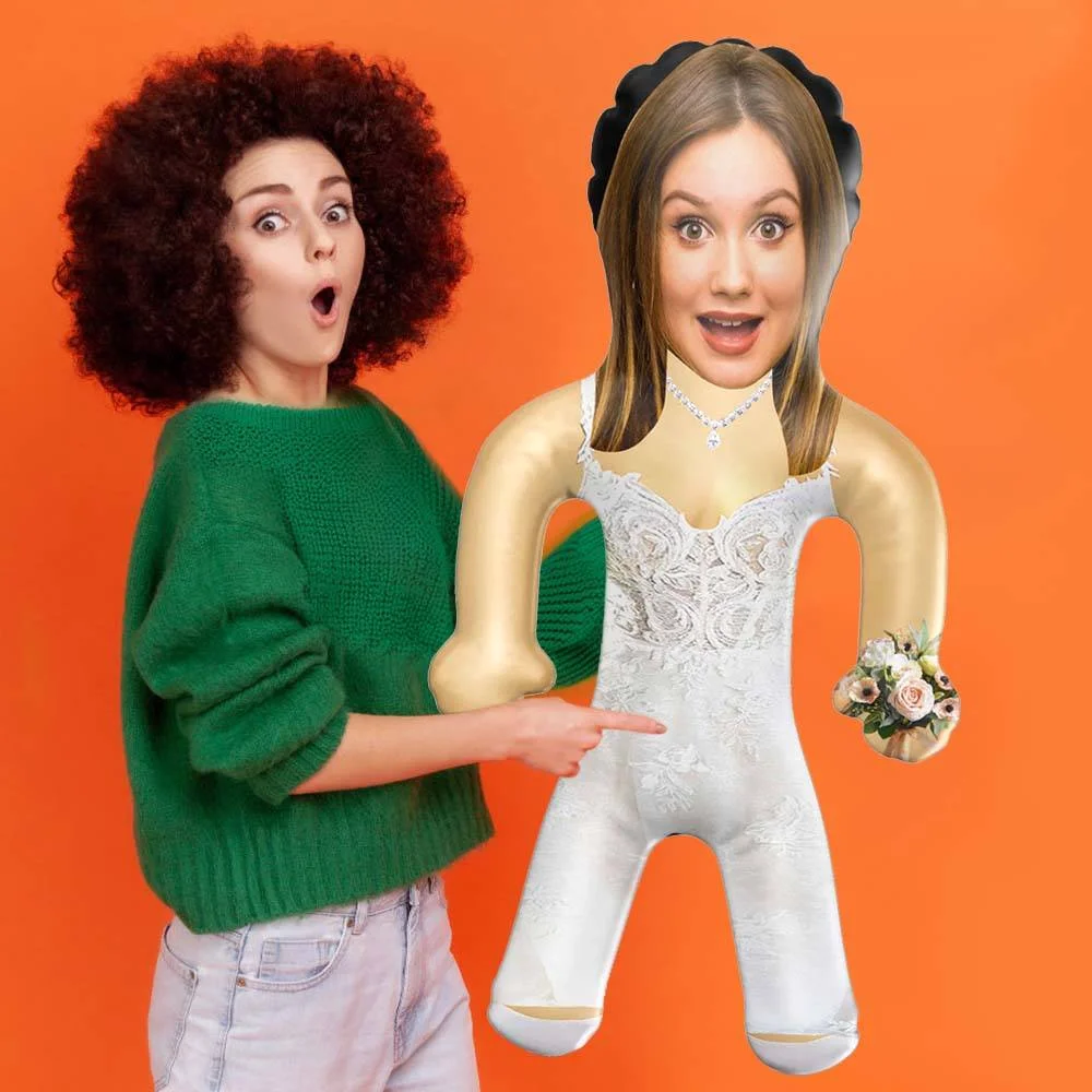 Woman in a green sweater points at a large bridal puppet wearing a wedding dress against an orange background, with surprised expression.
