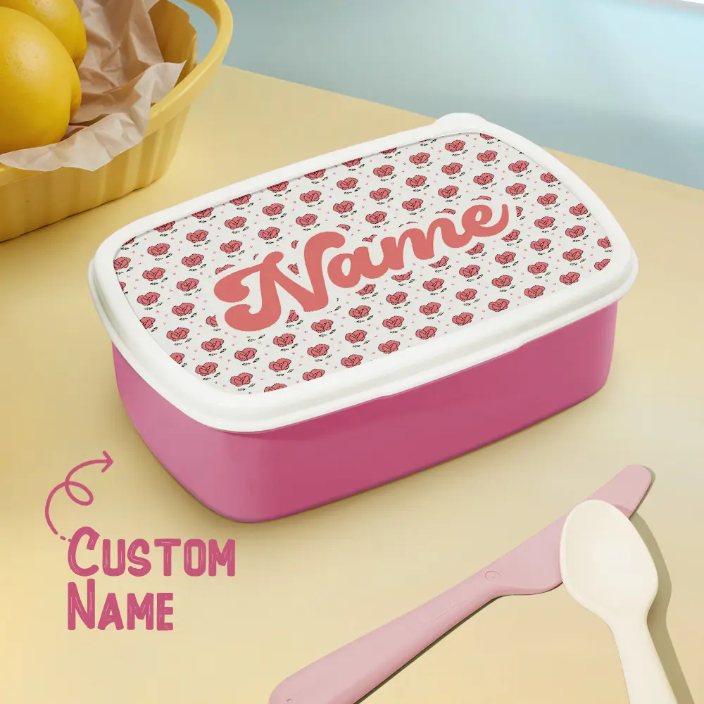 Pink lunchbox with a white lid covered in tiny pink hearts and the word 'Name' in pink script, indicating a customizable name. Nearby are a pink butter knife and spoon and a basket of lemons.