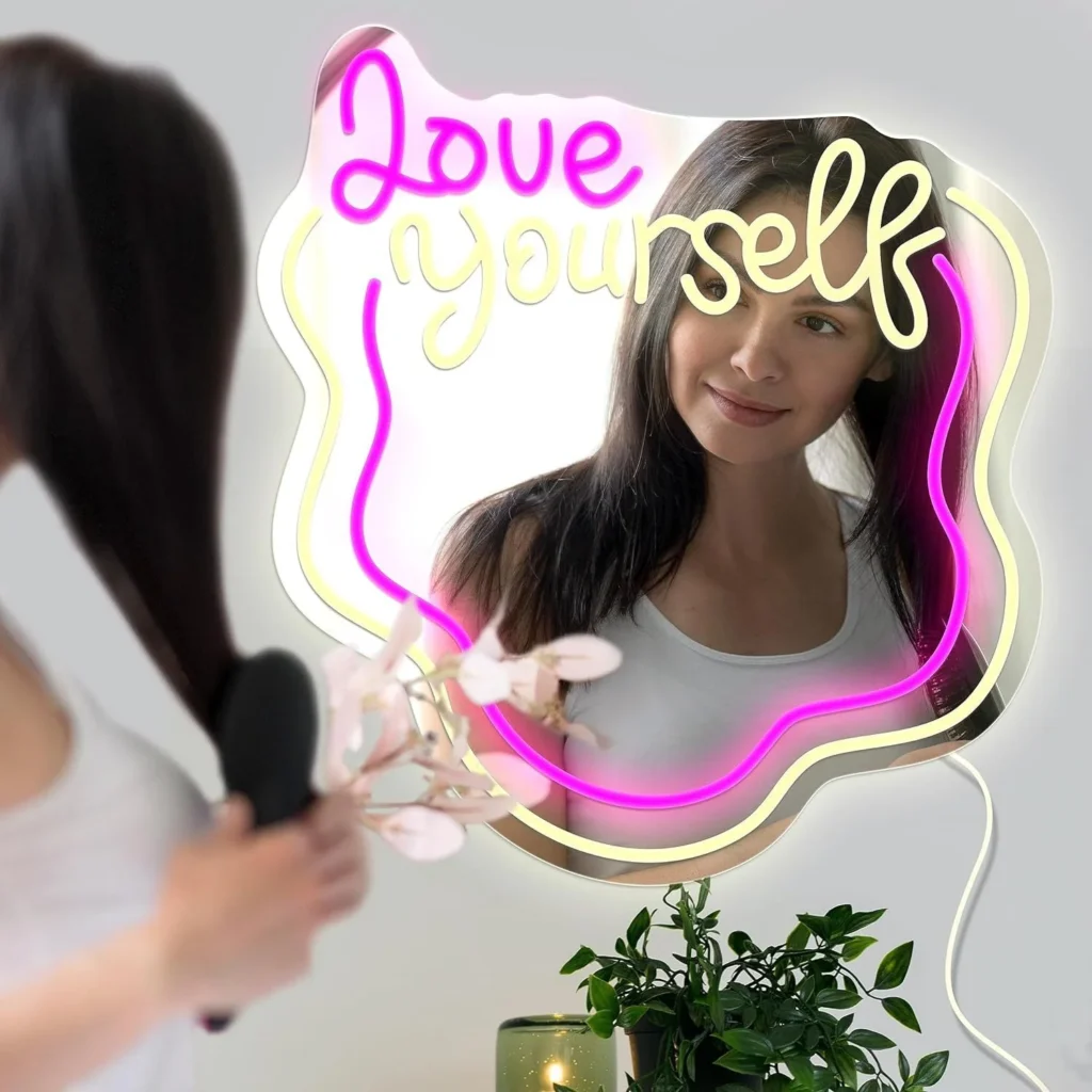 Woman admiring her reflection in a neon-framed mirror that reads 'Love Yourself' in bright pink and yellow lights (self-care scene).