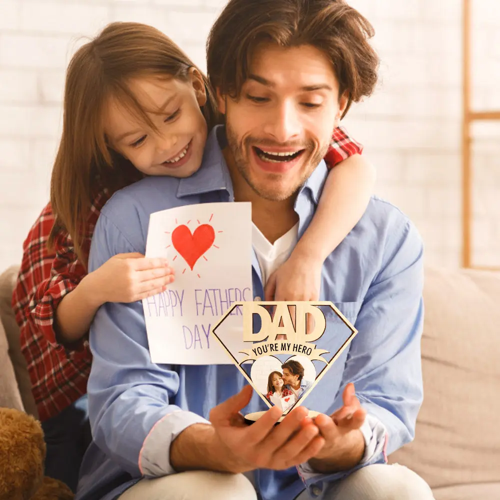 A father wearing a blue shirt smiles as a girl hugs him from behind, holding a handmade card with a red heart and Happy Father's Day message next to a superhero-style Dad sign.
