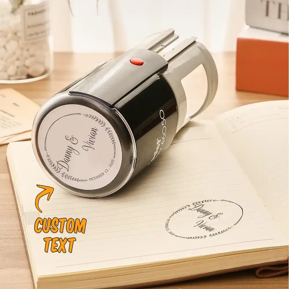 Gray self-inking stamp resting on an open notebook, showing a customizable text stamp and a circular imprint reading 'Dany & Nadia'.