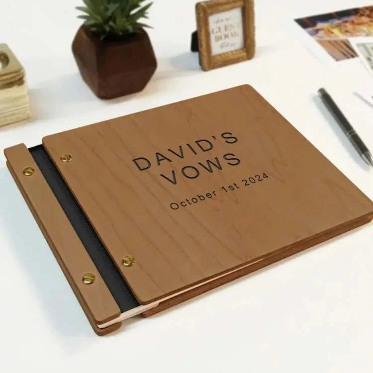 Close-up of a wooden vow book reading 'DAVID'S VOWS' with date October 1st 2024 on a white desk, plant and frame in the background.