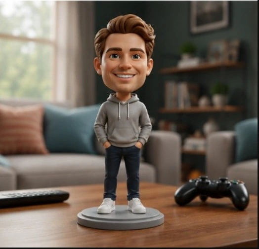 Bobblehead figurine of a smiling man in a gray hoodie and jeans standing on a round base on a coffee table, with a game controller nearby in a living room.