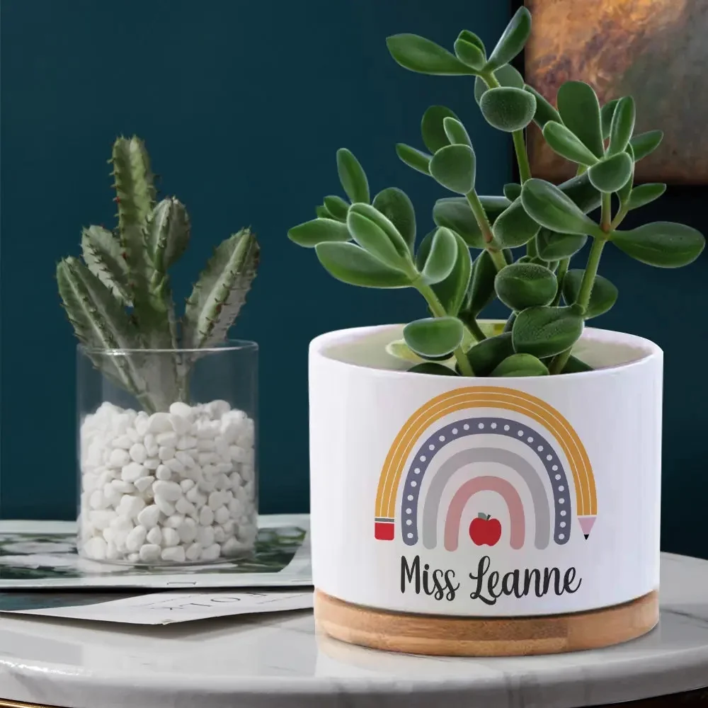 White ceramic pot with a colorful rainbow design and 'Miss Leanne' text, containing a green succulent on a wooden base.