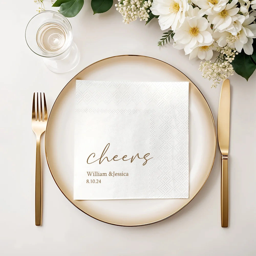 Wedding reception place card on a gold-rimmed plate with 'cheers' and the names William & Jessica, dated 8.10.24, beside white flowers and gold utensils.