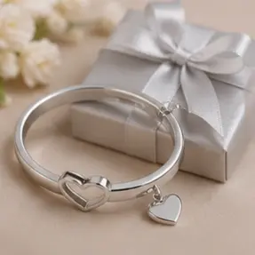 Silver bracelet with a heart charm, placed beside a white gift box tied with a satin ribbon.
