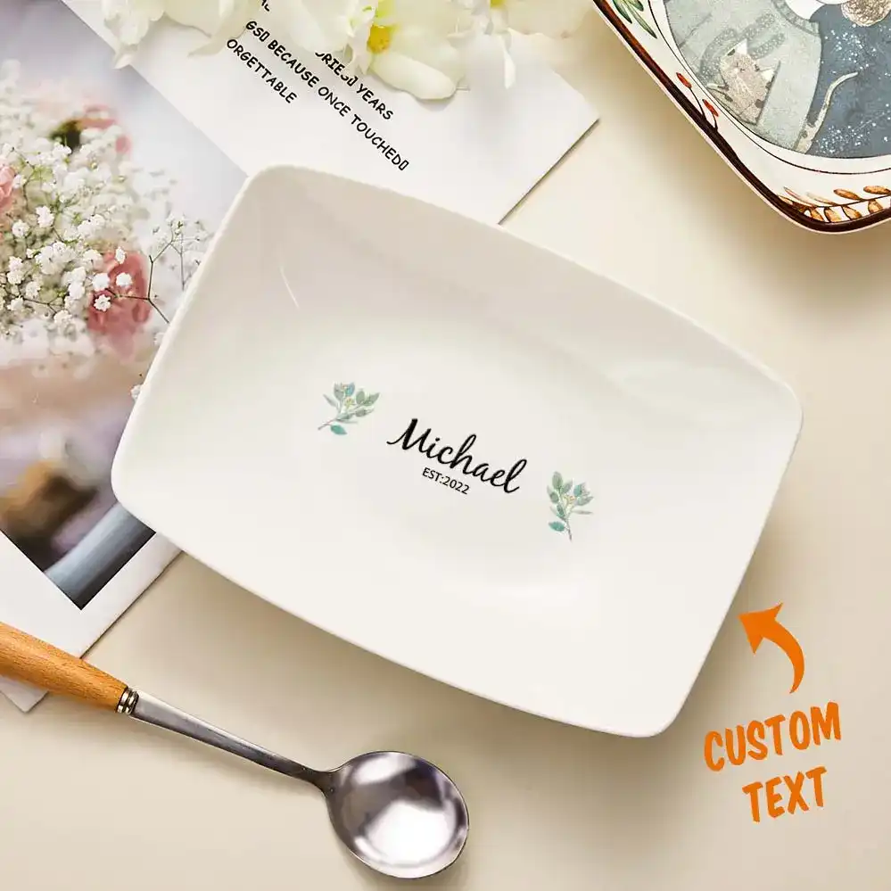 White rectangular plate with the script name "Michael" and "EST-2022" flanked by small floral accents, illustrating a custom text design on tableware.