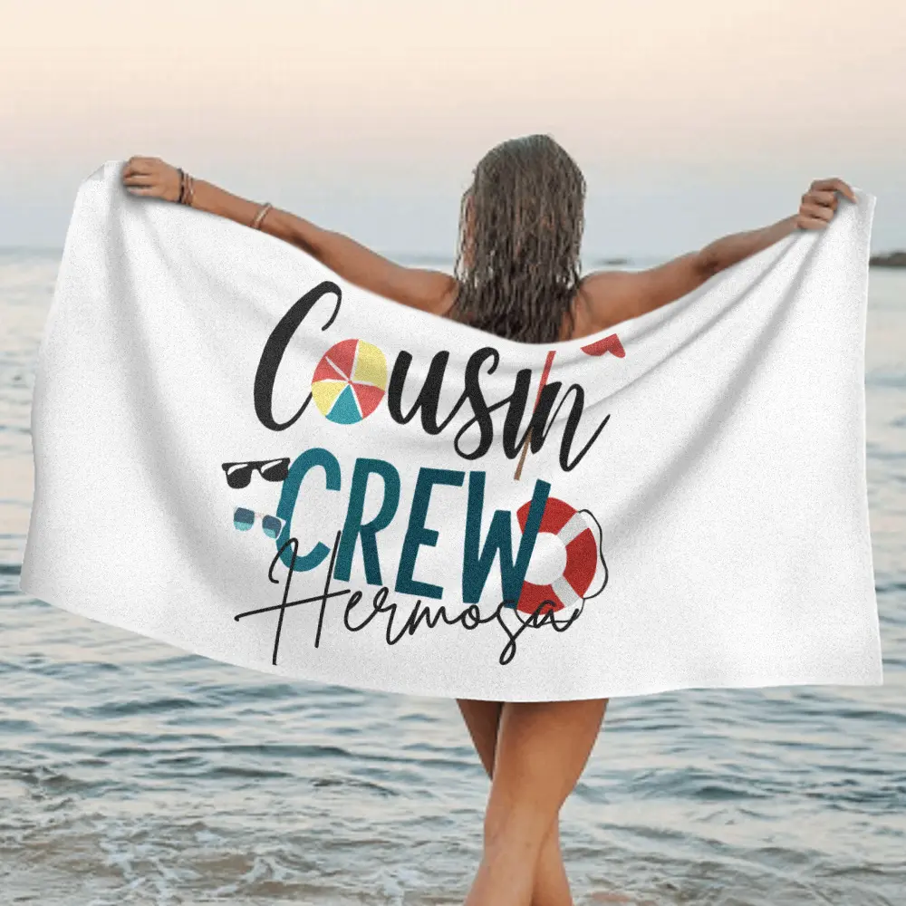 Person standing in shallow ocean water at sunset, holding a white towel that reads 'Cousin Crew Hermosa' with colorful icons.
