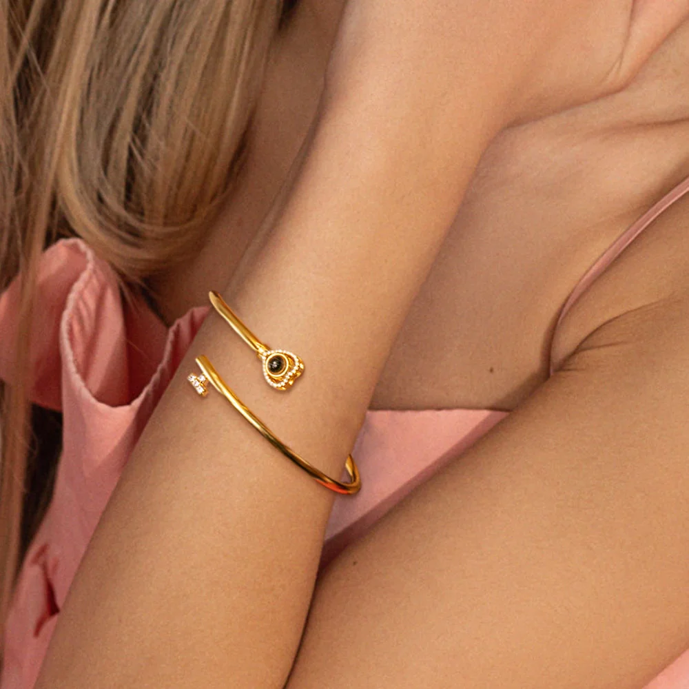 Gold open-cuff bracelet on a wrist, featuring a circular charm with a black center and a small diamond charm against pink fabric.