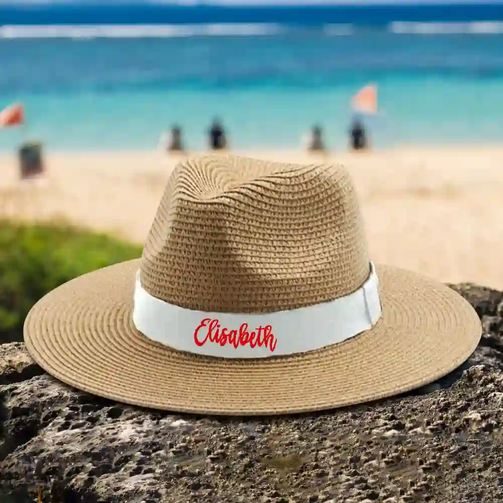 Straw sun hat with white band and red name 'Elizabeth' resting on a rocky beach with the ocean in the background.