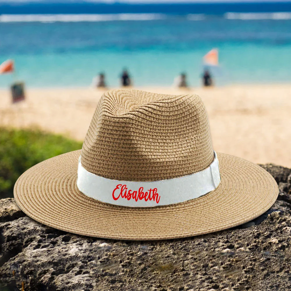 Straw sunhat with a white band and red 'Elisabeth' text on a rocky beach with the blue sea in the background.