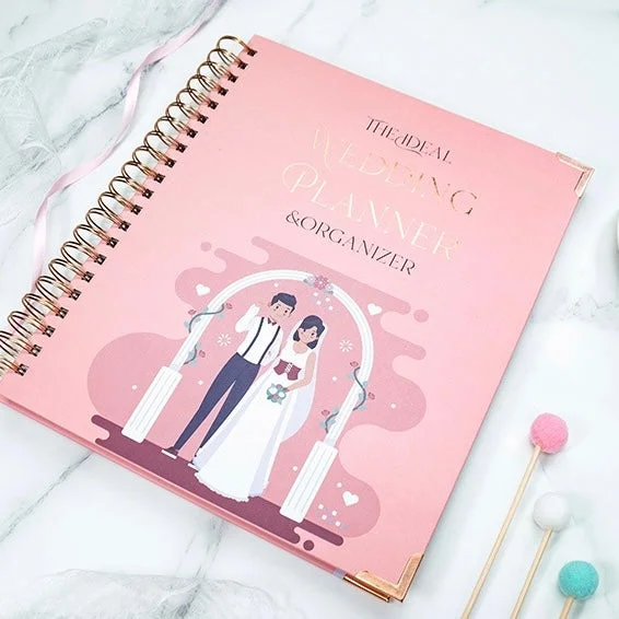 Pink wedding planner notebook with a bride and groom under a decorative arch on the cover, gold spiral binding.