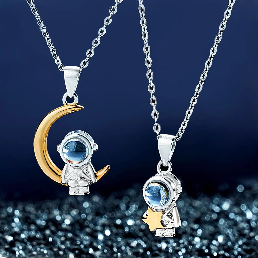 Two space-themed pendant necklaces featuring astronaut charms with blue gemstone helmets, one perched on a gold crescent moon.