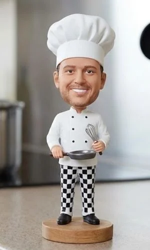 Bobblehead chef figurine in white uniform and checkered pants, holding a whisk and frying pan on a wooden base.