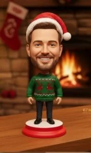 Bobblehead figurine with a man's head, wearing a Santa hat and green Christmas sweater, standing on a red base by a fireplace.