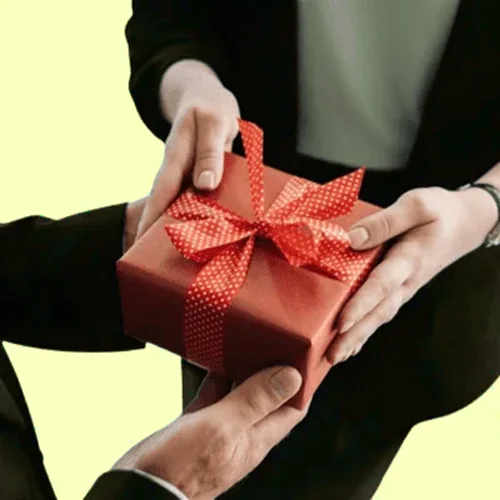 Two people exchanging a neatly wrapped red gift with a ribbon against a pale yellow background.