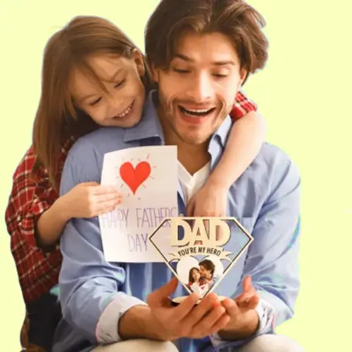Father and daughter smiling together while holding a Father’s Day card with a heart and a 'Dad, you're my hero' sign.