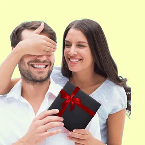 Smiling couple; woman hands man a black gift box with a red ribbon as he playfully covers his eyes.